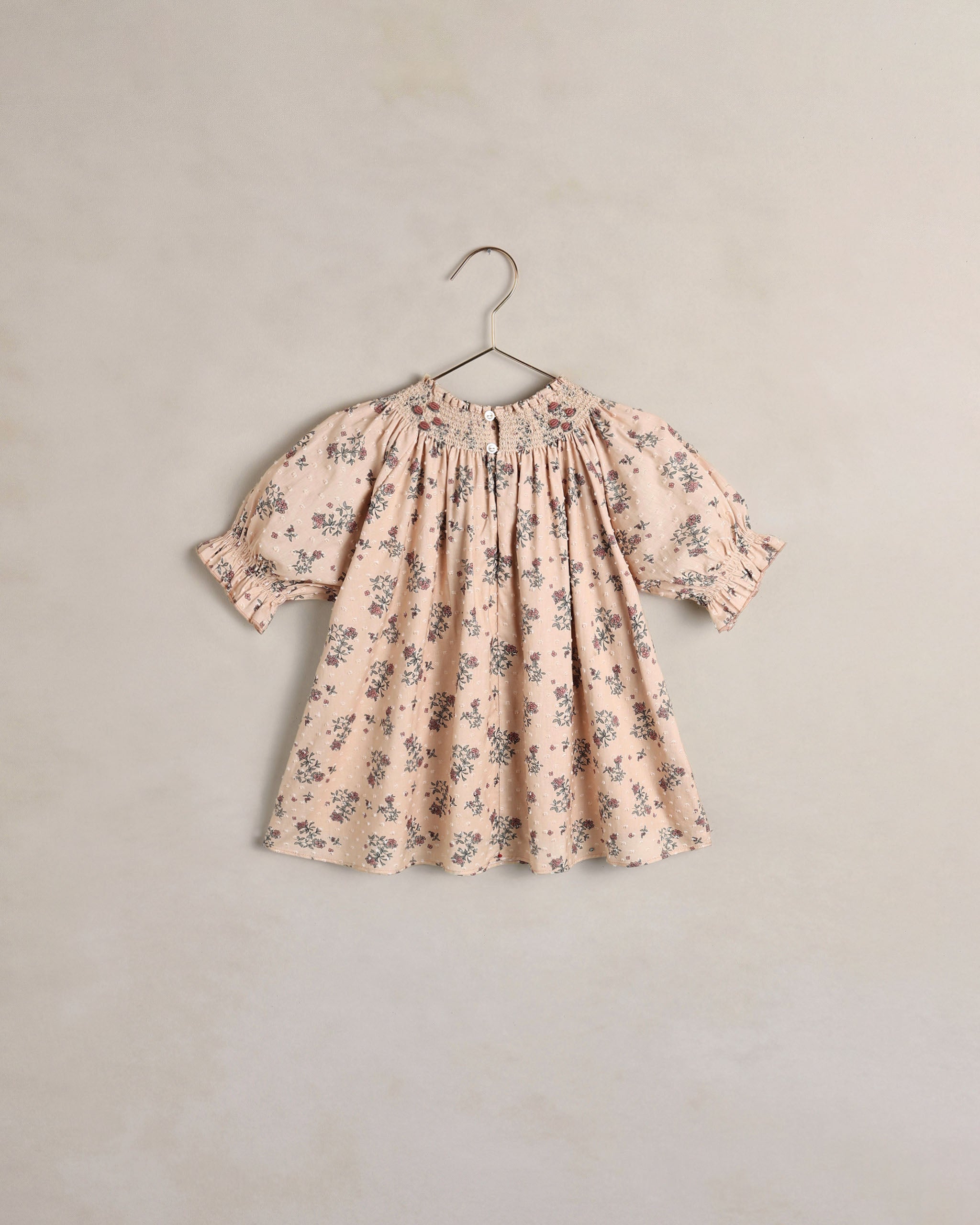 A children's short sleeve dress with a smocked bodice and ruffled sleeves, featuring a delicate floral print in muted blue and green on a petal pink. The dress is shown hanging on a simple metal hanger against a textured, warm beige wall.