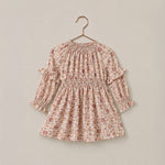 A long-sleeve baby dress with a delicate pink and brown floral print, featuring a smocked waist and ruffled sleeves, hanging on a wooden hanger against a solid beige background.