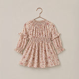A long-sleeve baby dress with a delicate pink and brown floral print, featuring a smocked waist and ruffled sleeves, hanging on a wooden hanger against a solid beige background.