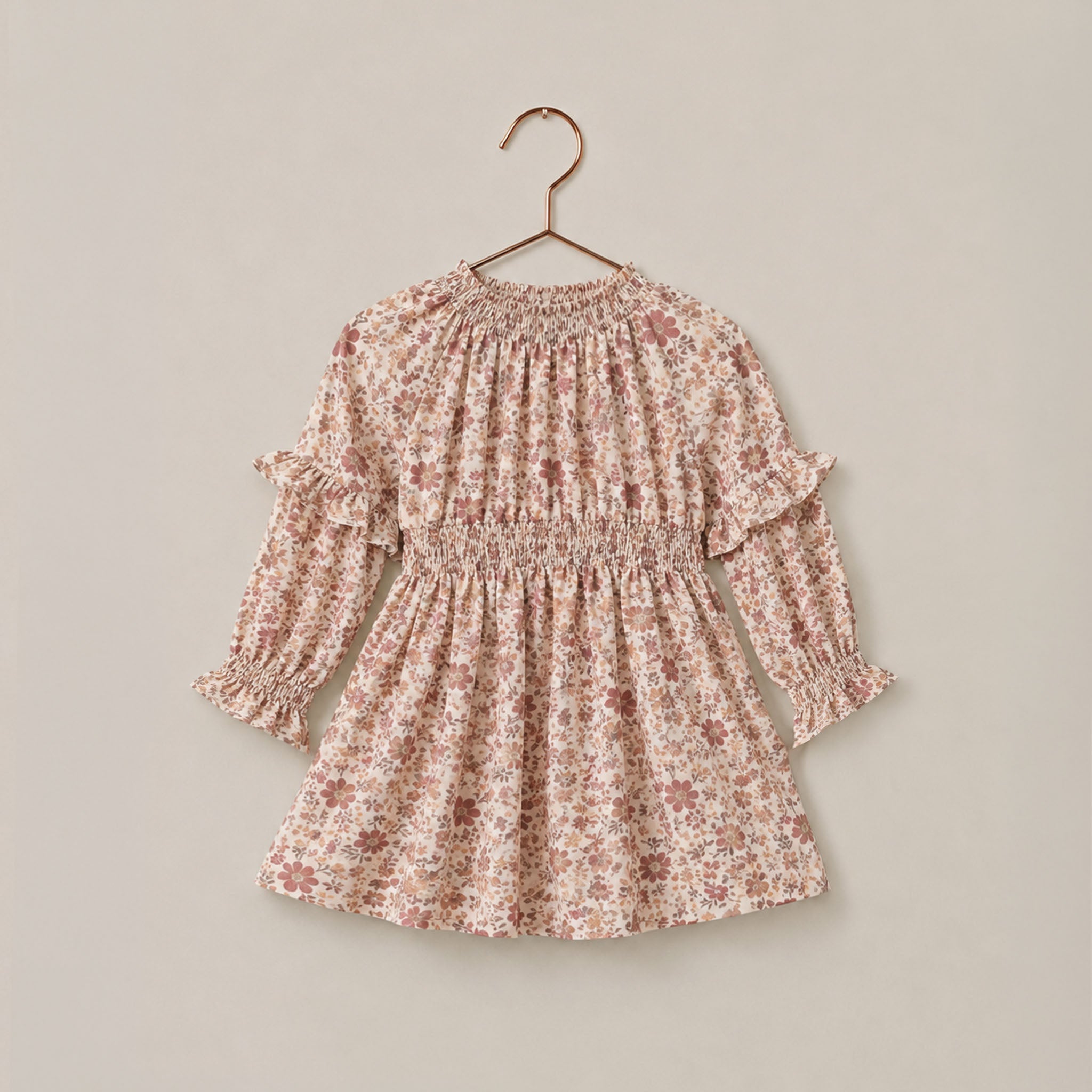 A long-sleeve baby dress with a delicate pink and brown floral print, featuring a smocked waist and ruffled sleeves, hanging on a wooden hanger against a solid beige background.