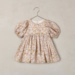 A toddler girl's dress with puff sleeves and a gathered smocked bodice, featuring a delicate beige and brown floral toile pattern, hanging on a metal hanger against a textured beige wall.