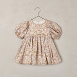 A toddler girl's dress with puff sleeves and a gathered smocked bodice, featuring a delicate beige and brown floral toile pattern, hanging on a metal hanger against a textured beige wall.