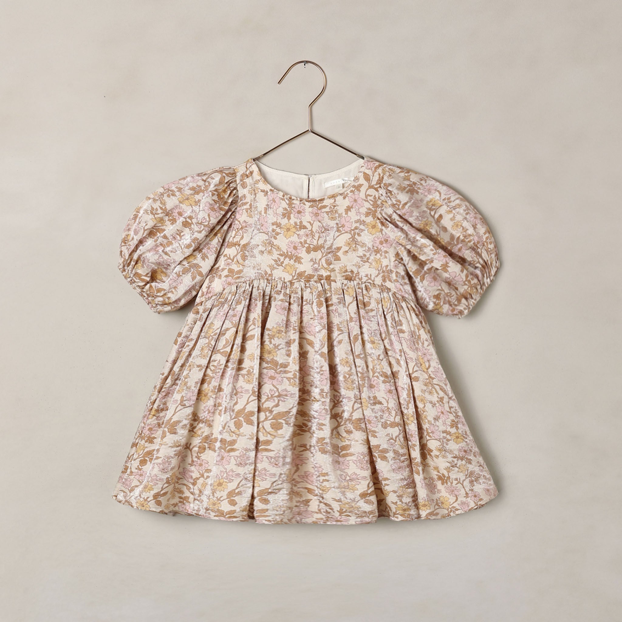 A toddler girl's dress with puff sleeves and a gathered smocked bodice, featuring a delicate beige and brown floral toile pattern, hanging on a metal hanger against a textured beige wall.