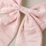 A close-up of a large light pink bow made from a soft, slightly textured fabric, set against a neutral beige background.
