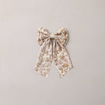 A bow-shaped fabric hair accessory with a delicate floral pattern in muted beige, brown, and soft pink tones, centered on a plain light gray background.