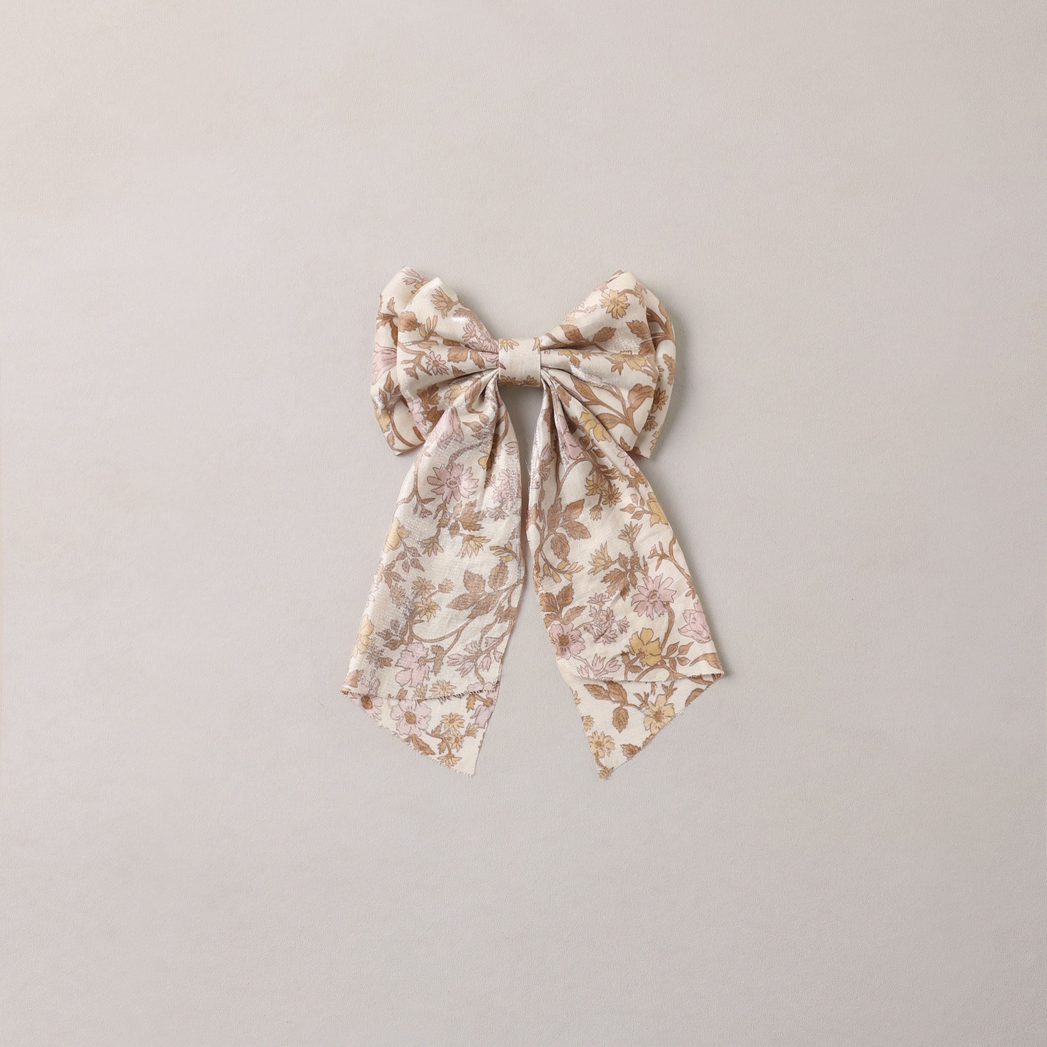 A bow-shaped fabric hair accessory with a delicate floral pattern in muted beige, brown, and soft pink tones, centered on a plain light gray background.