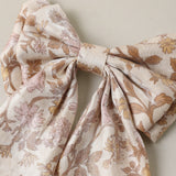 A large fabric bow featuring a delicate floral and leaf pattern in muted brown and dusty pink on a warm cream background, photographed against a neutral beige wall.