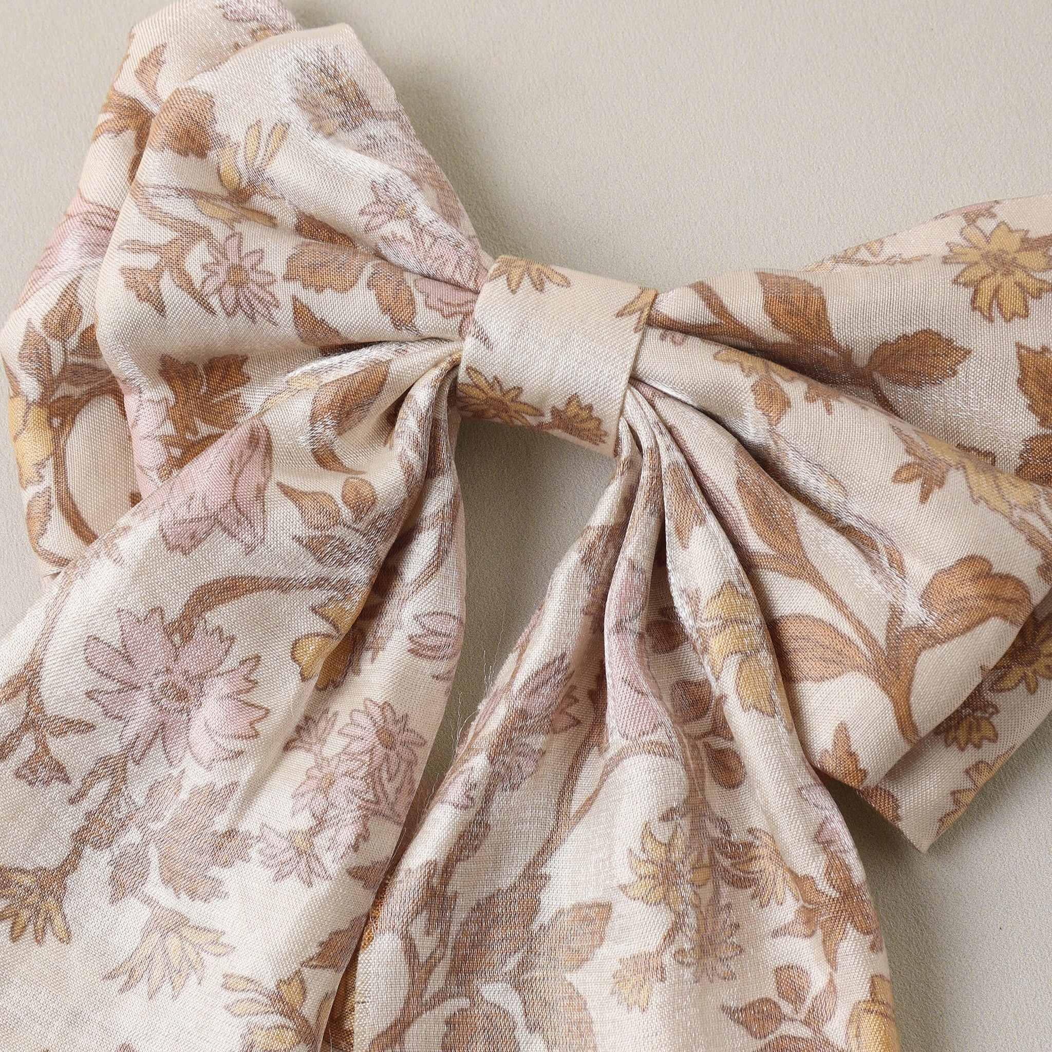 A large fabric bow featuring a delicate floral and leaf pattern in muted brown and dusty pink on a warm cream background, photographed against a neutral beige wall.