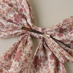 A close-up of a sheer floral hair bow with a gold metal claw clip, featuring a pattern of dusty pink and brown flowers on a light beige background.