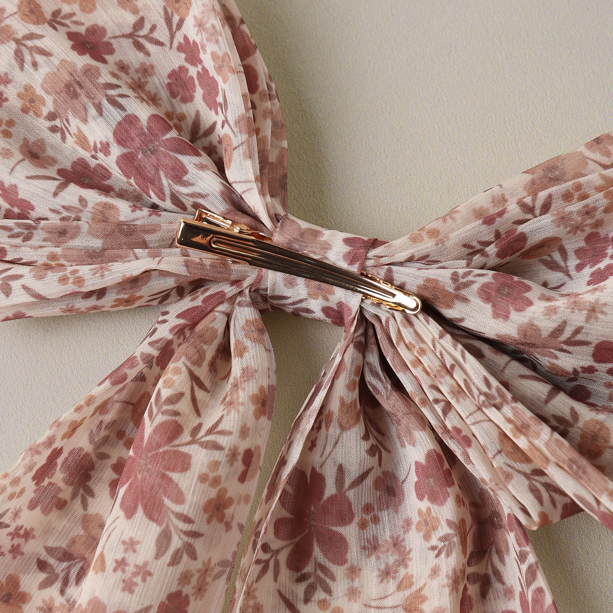 A close-up of a sheer floral hair bow with a gold metal claw clip, featuring a pattern of dusty pink and brown flowers on a light beige background.