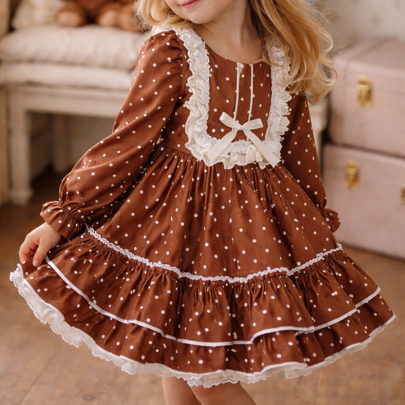 A young girl with blonde hair wearing a brown long-sleeve dress with white polka dots, white lace trim, and a white bow at the neckline, standing in a cozy room with wooden floors and a teddy bear in the background.