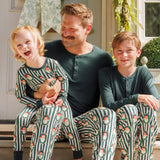 A father and two sons wearing matching green and white striped holiday pajamas with a Santa Claus pattern, sitting together on the front steps of a house.