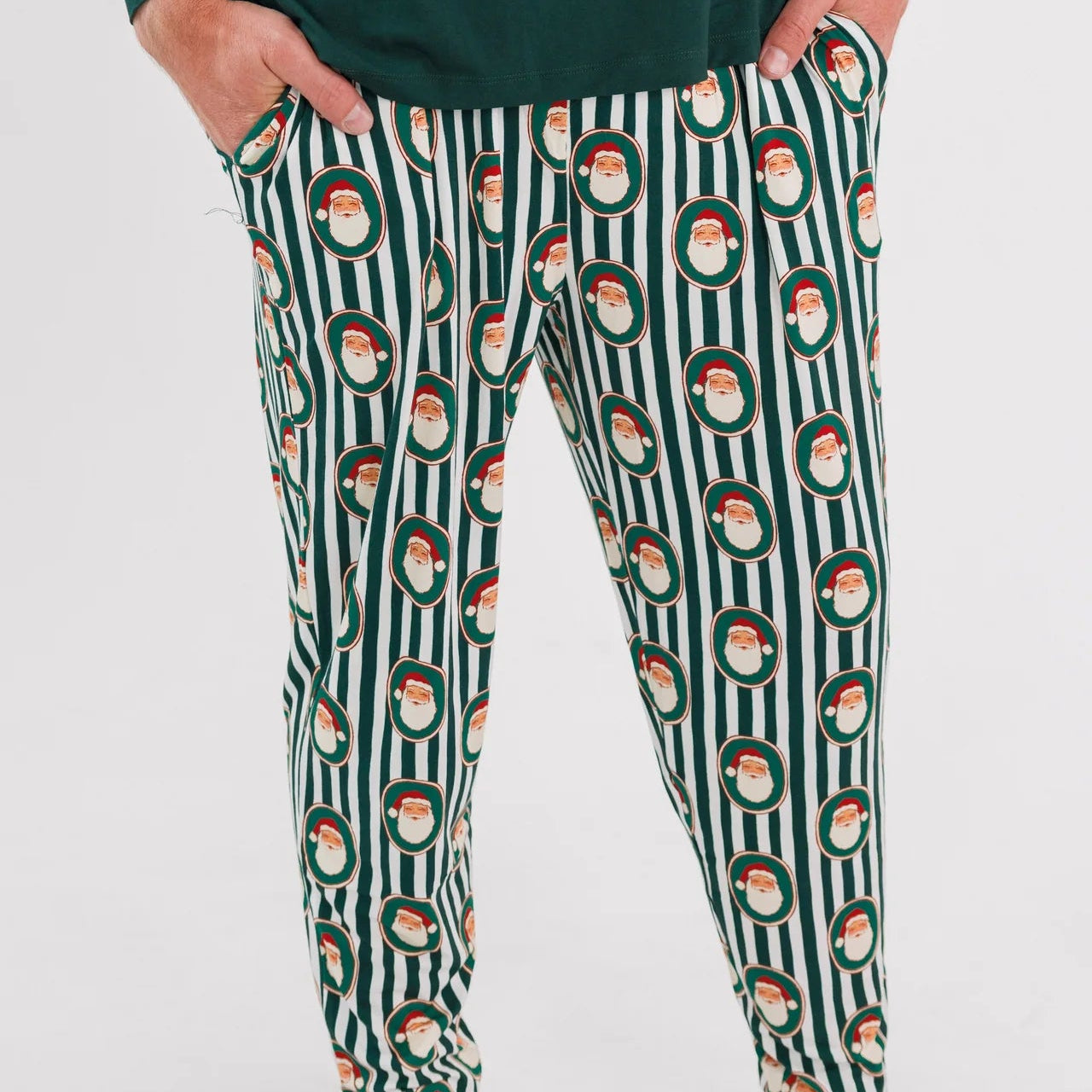 A person wearing dark green pajama pants with a vertical white stripe pattern and a repeating Santa Claus face motif, paired with a dark green long-sleeve top. The photo is cropped from the chest down against a plain white studio background.