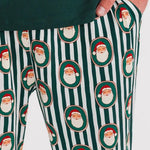 A close-up of a person wearing holiday-themed lounge pants with a green and white vertical stripe pattern and repeated Santa Claus face motifs, paired with a dark green top.