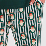 A close-up of a person wearing holiday-themed lounge pants with a green and white vertical stripe pattern and repeated Santa Claus face motifs, paired with a dark green top.