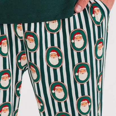 A close-up of a person wearing holiday-themed lounge pants with a green and white vertical stripe pattern and repeated Santa Claus face motifs, paired with a dark green top.