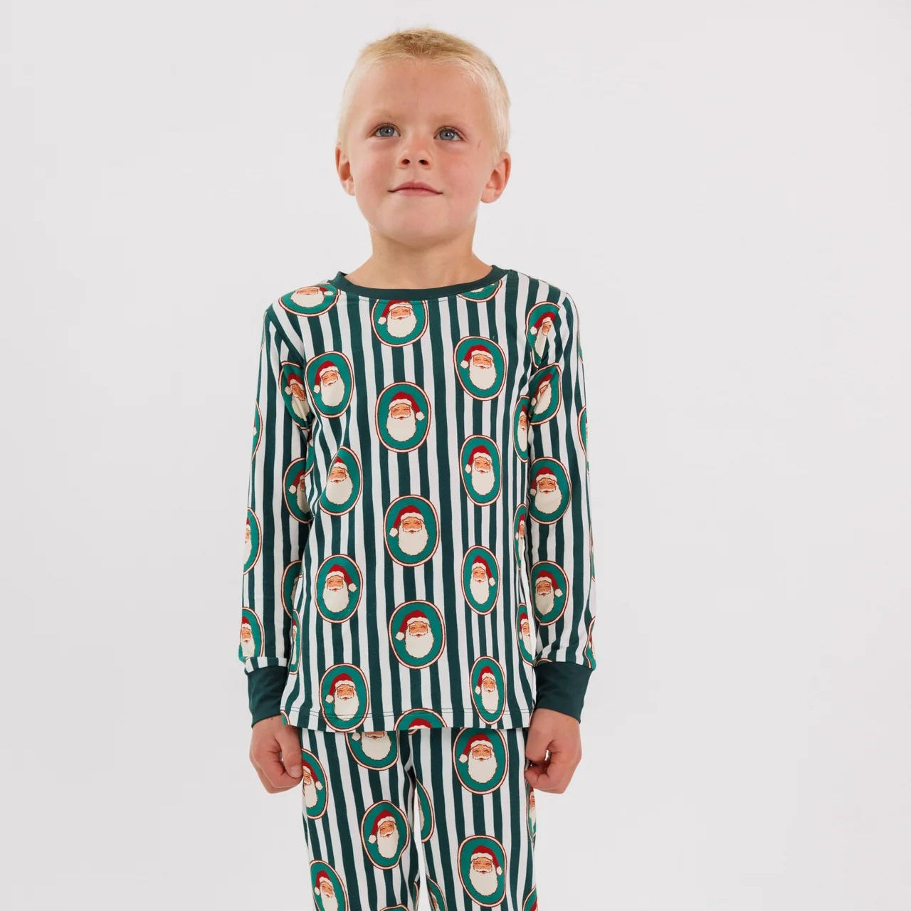 A young child standing against a light gray studio background, wearing a long-sleeve pajama set with dark green and white vertical stripes and a repeating Santa Claus face pattern.