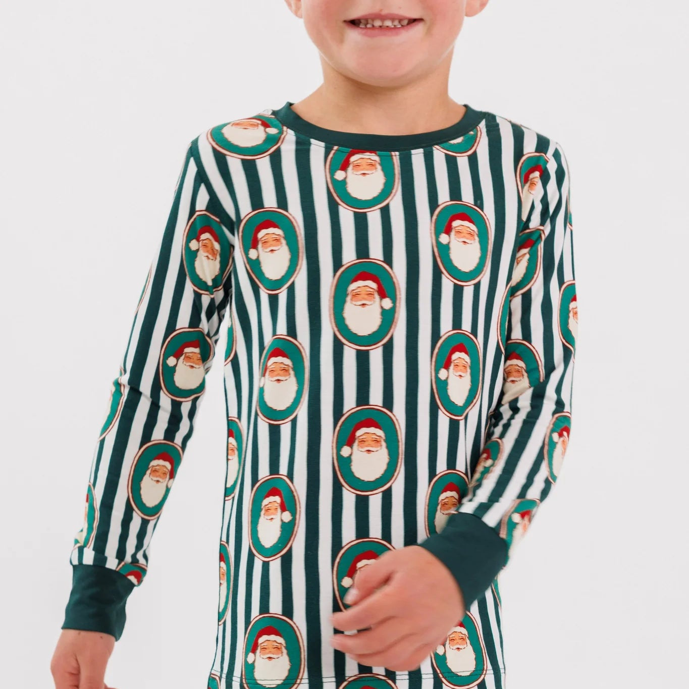 A child wearing a long-sleeve and matching pajama set with dark green and white vertical stripes and an allover Santa Claus face print, shown against a light gray studio background.