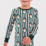 A child wearing a long-sleeve and matching pajama set with dark green and white vertical stripes and an allover Santa Claus face print, shown against a light gray studio background.