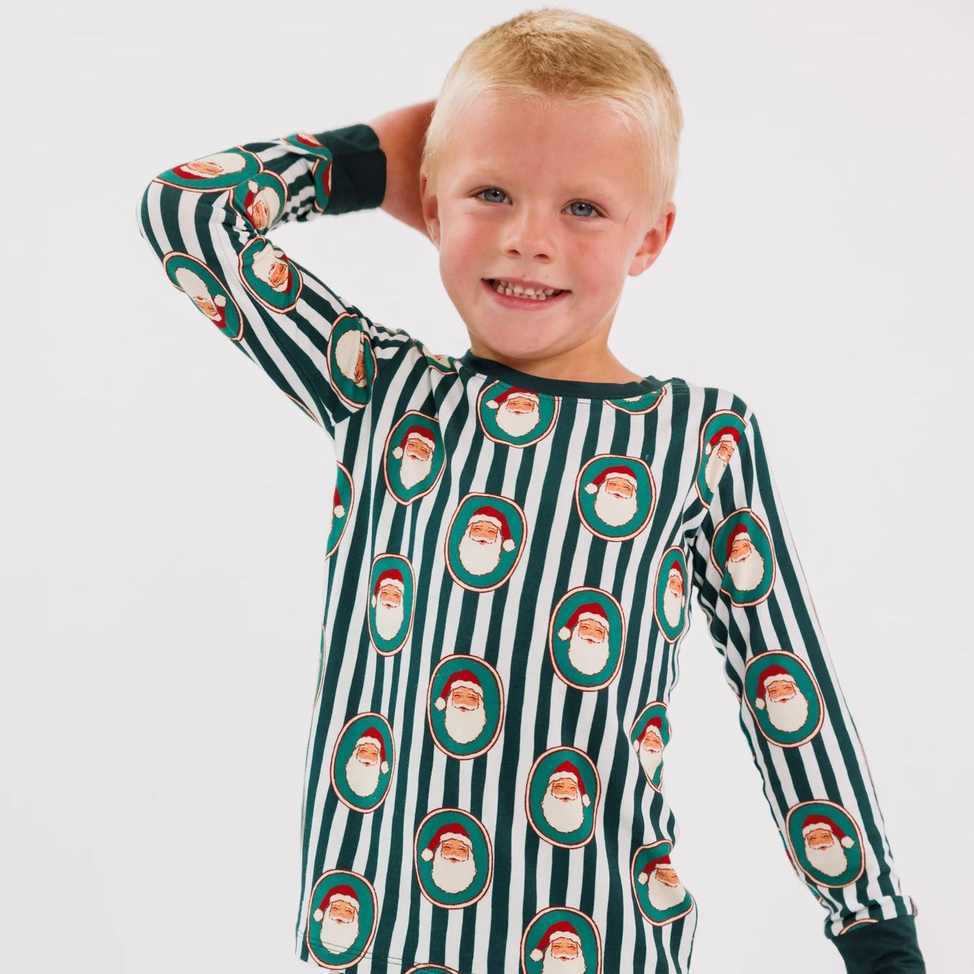 A young child wearing a long-sleeve pajama set with dark green and white vertical stripes and an all-over Santa Claus face print, posed against a plain light gray studio background.