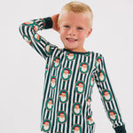 A young child wearing a long-sleeve pajama set with dark green and white vertical stripes and an all-over Santa Claus face print, posed against a plain light gray studio background.