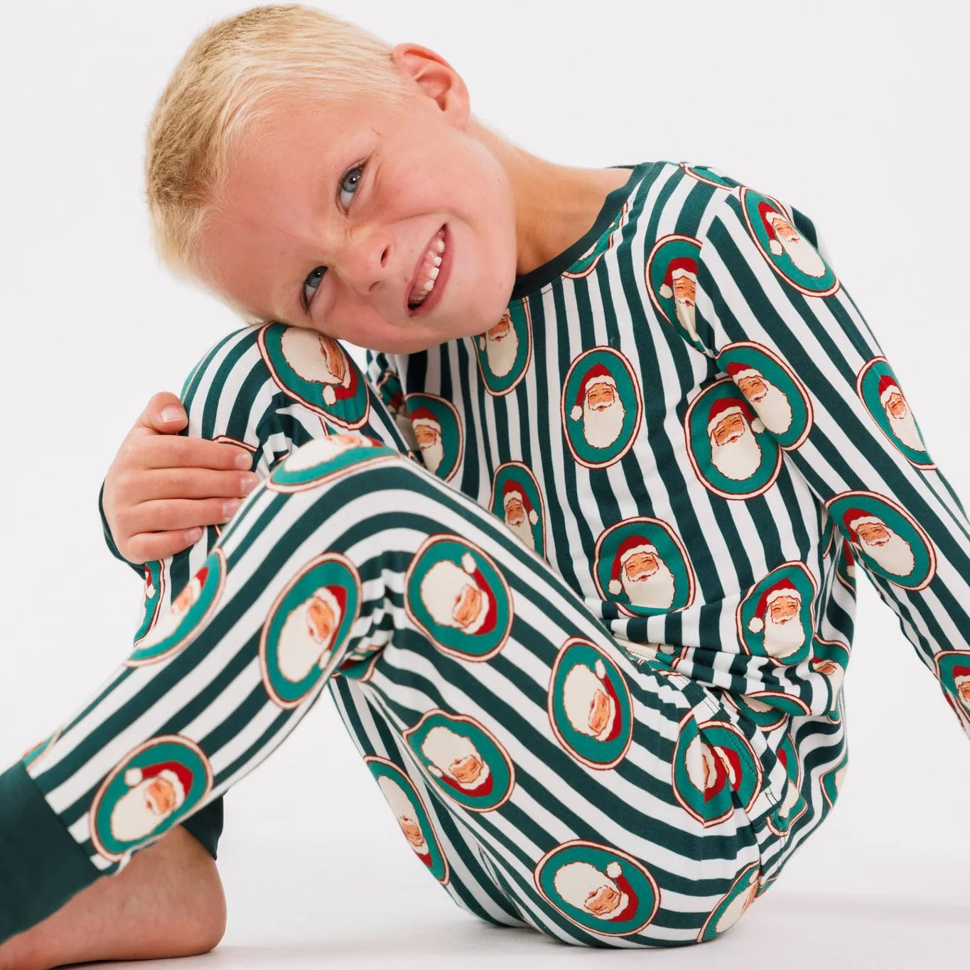 A young blonde boy sitting on a white studio floor, wearing a long-sleeve pajama set with dark green and white stripes and a repeating pattern of Santa Claus faces inside red and green circular frames.