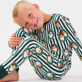 A young blonde boy sitting on a white studio floor, wearing a long-sleeve pajama set with dark green and white stripes and a repeating pattern of Santa Claus faces inside red and green circular frames.
