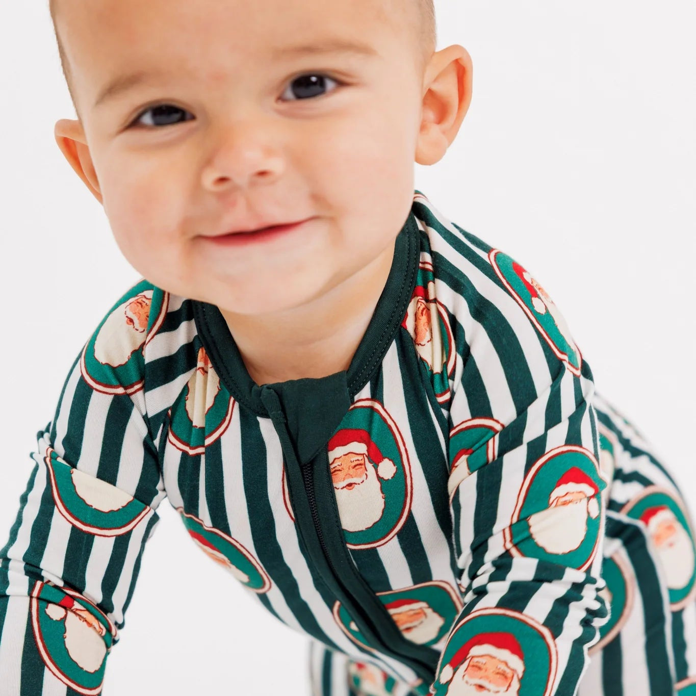 A baby crawling on a white studio floor wearing a green and white striped long-sleeve pajama with a Santa Claus print.