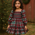 A young girl with long dark curly hair stands outdoors wearing a festive holiday plaid dress. The dress features a smocked bodice, long puff sleeves, and a tiered ruffle skirt in a red, green, and white tartan pattern.