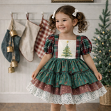 A young girl with curly brown hair wearing a festive Christmas tiered dress, standing in a decorated room with stockings, a Christmas tree, and wooden floors.