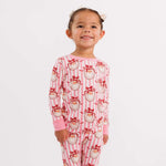 A young girl stands barefoot against a plain white studio background wearing a one-piece pajama set with pink and white vertical stripes and a repeating Santa face print in red, white, and black.
