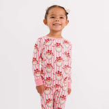 A young girl stands barefoot against a plain white studio background wearing a one-piece pajama set with pink and white vertical stripes and a repeating Santa face print in red, white, and black.
