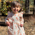 A young child wearing a white long-sleeve dress with a red and orange butterfly print, standing outdoors in a wooded setting.