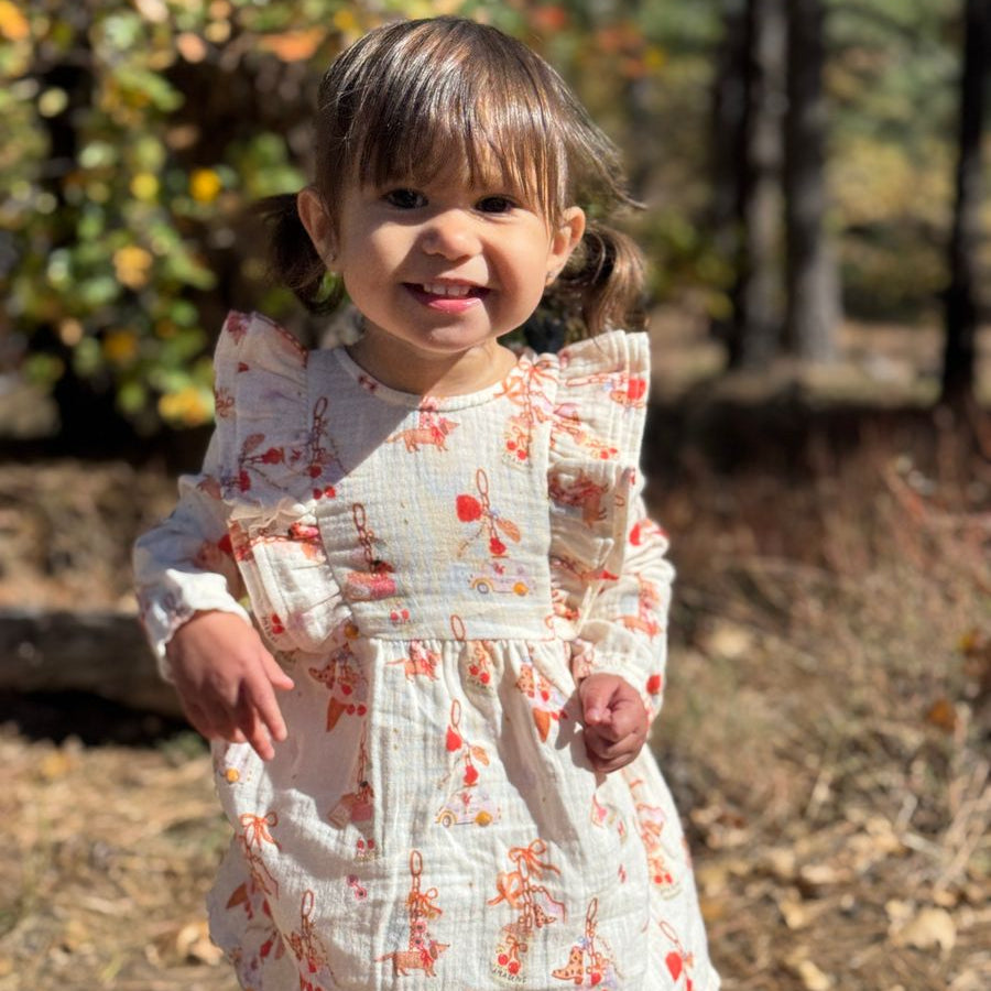 A young child wearing a white long-sleeve dress with a red and orange butterfly print, standing outdoors in a wooded setting.