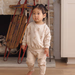 A toddler girl stands barefoot in a cozy indoor setting, wearing a light beige two-piece pajama set with small mitten print. The background features a rustic stone fireplace with a wooden sled and a light-colored cabinet.