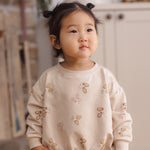 A toddler girl wearing a cream-colored sweatshirt and matching pants set, decorated with a pattern of different colored mitten, standing in a softly lit indoor room.