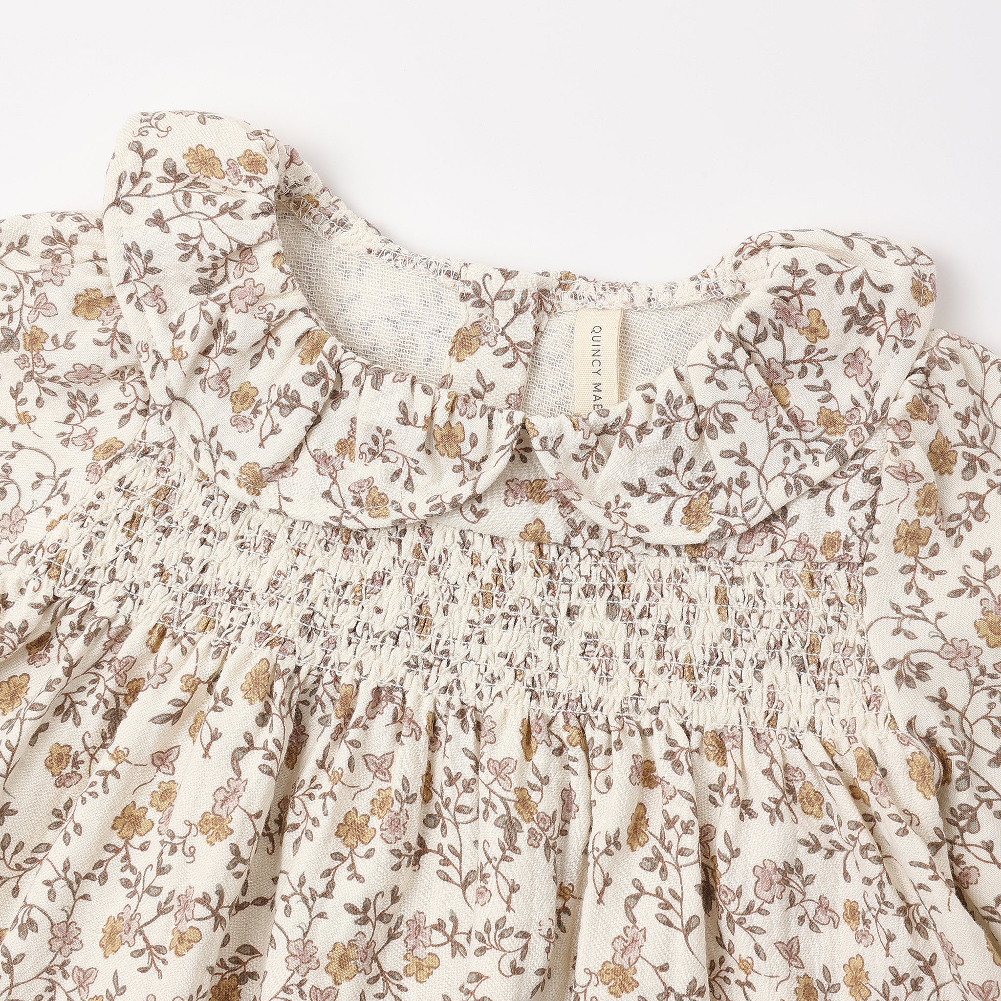 Close-up of a cream-colored children's dress with a brown and muted yellow floral print, featuring a smocked bodice and ruffled collar.