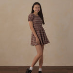 A young woman modeling a short puff-sleeve plaid dress in brown and beige tones, paired with white socks and black loafers, standing against a warm beige wall and dark wood floor.