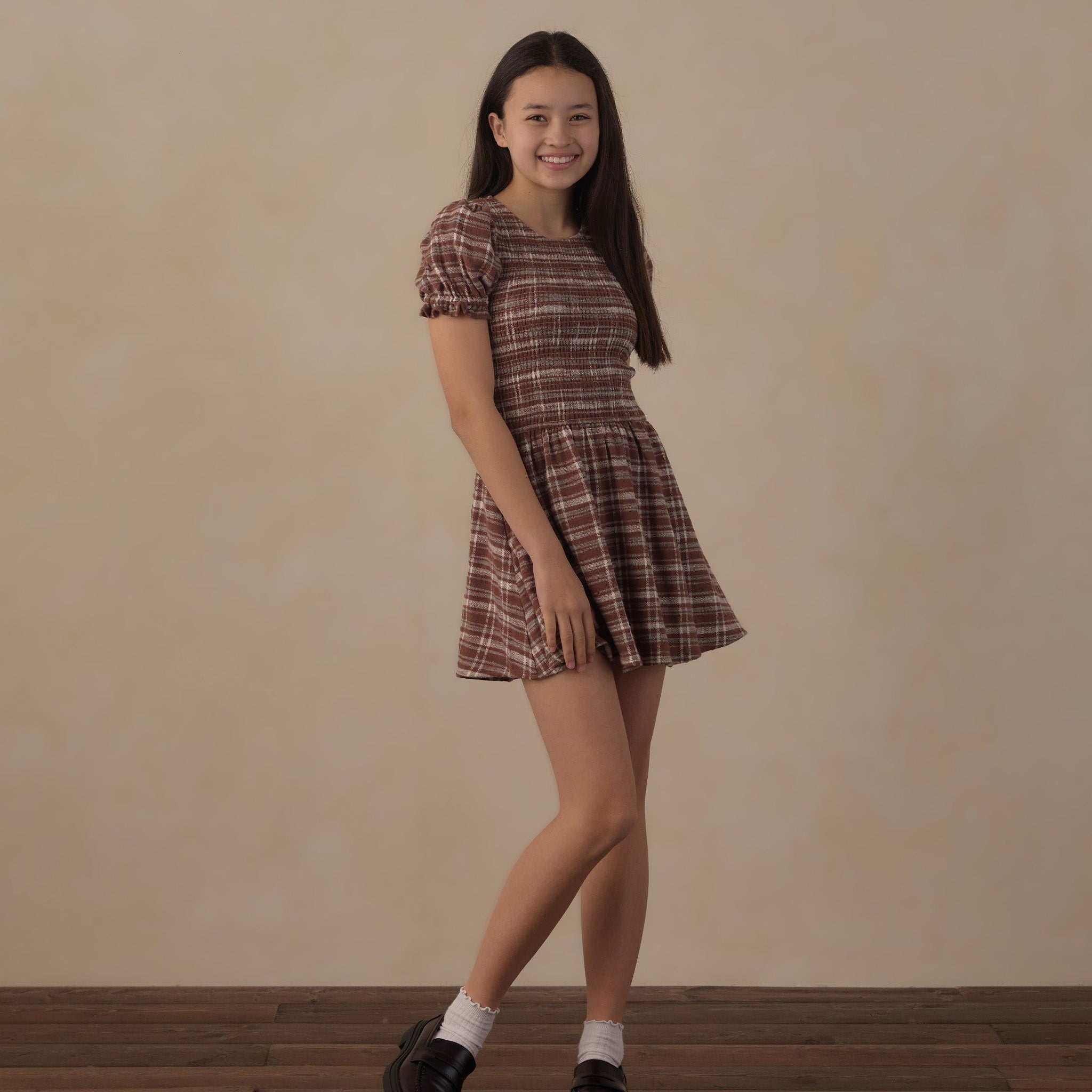 A young woman modeling a short puff-sleeve plaid dress in brown and beige tones, paired with white socks and black loafers, standing against a warm beige wall and dark wood floor.