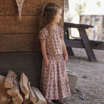 A young girl with curly brown hair stands outdoors in front of a rustic wooden wall, wearing a short-sleeve pink dress with a small floral print. A stack of firewood is visible at her left, and a wooden picnic table sits in the background.
