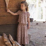 A young girl stands outdoors against a rustic wooden wall, wearing a short-sleeve smocked blouse and matching tiered skirt in a muted pink floral print. She holds a wooden sign, with stacked firewood and a dirt ground in the background.