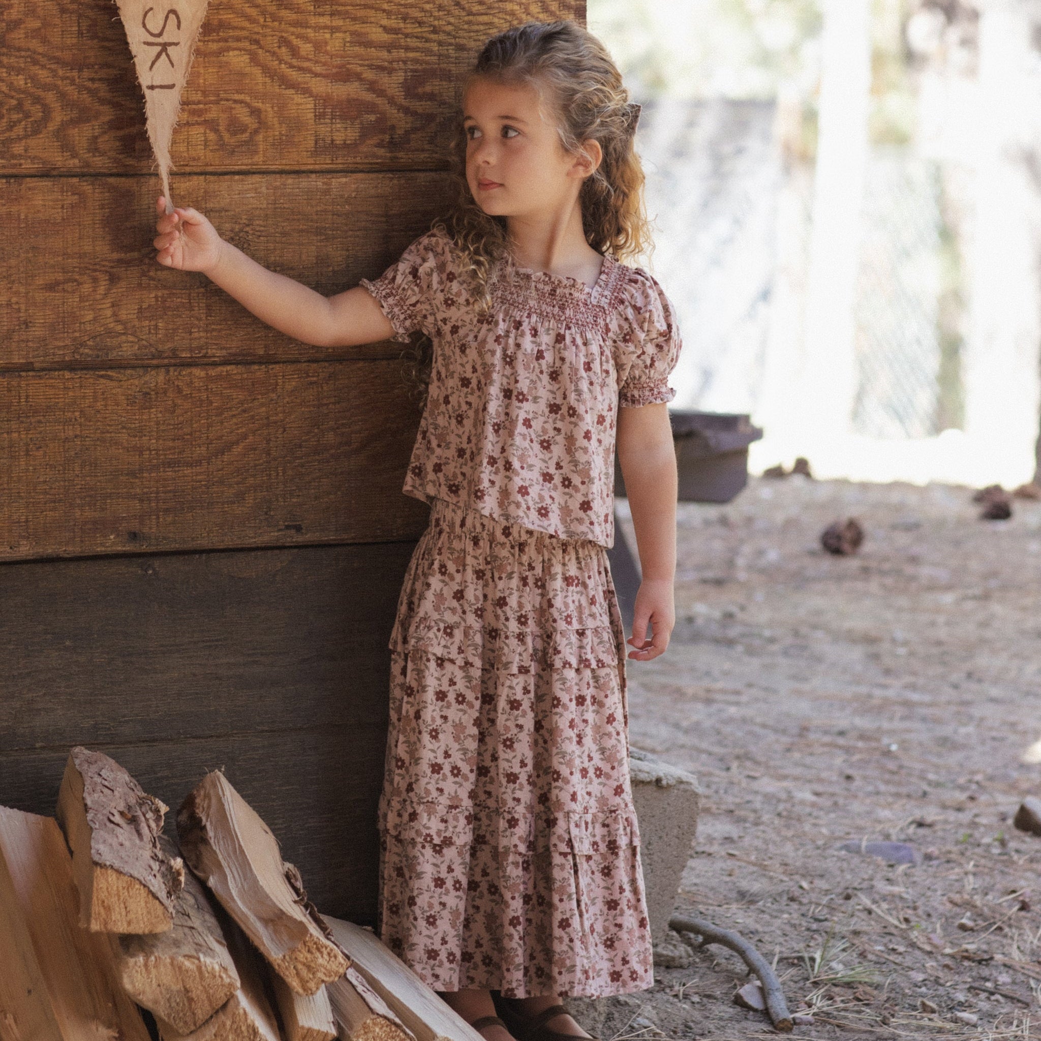 A young girl stands outdoors against a rustic wooden wall, wearing a short-sleeve smocked blouse and matching tiered skirt in a muted pink floral print. She holds a wooden sign, with stacked firewood and a dirt ground in the background.