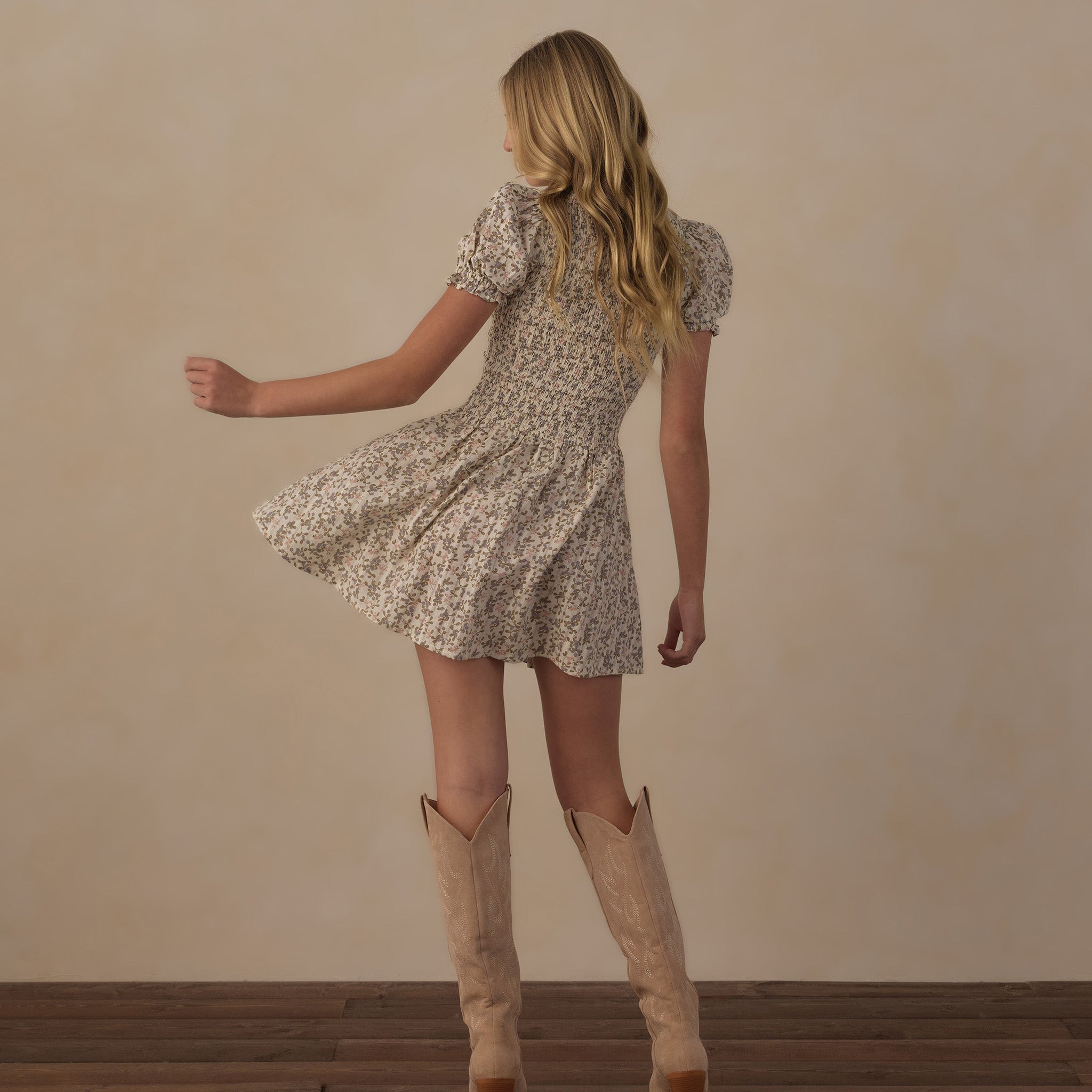 A woman is shown from behind wearing a short-sleeve cream and brown floral print mini dress and beige cowboy boots, standing on a dark wood floor against a textured beige wall.