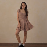 A young woman modeling a brown and beige gingham mini dress with puff sleeves, a square neckline, and a ruffled hem, paired with white socks and black leather shoes, standing against a textured beige wall and wooden floor.
