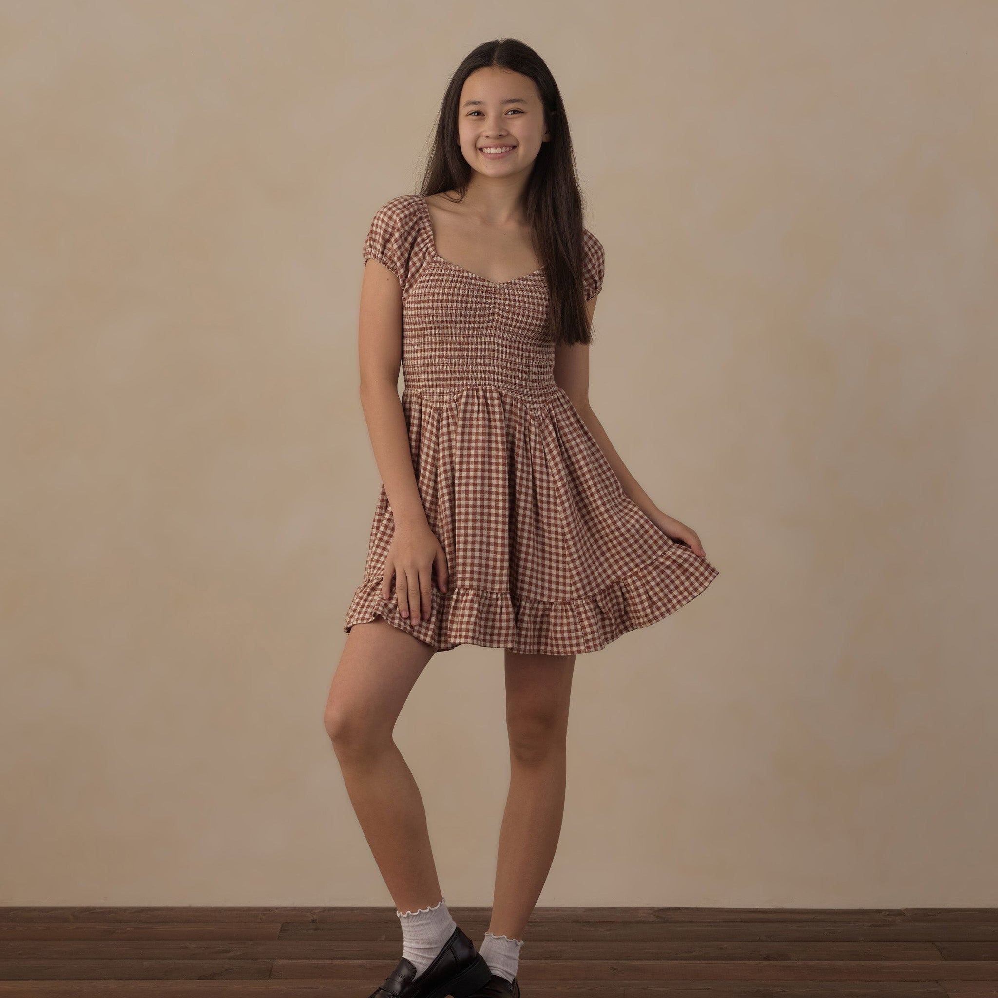 A young woman modeling a brown and beige gingham mini dress with puff sleeves, a square neckline, and a ruffled hem, paired with white socks and black leather shoes, standing against a textured beige wall and wooden floor.