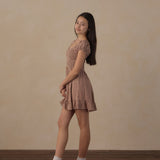 A young woman stands in profile wearing a short brown and beige gingham dress with puff sleeves, paired with white socks and black loafers, against a warm beige textured wall and dark wood floor.
