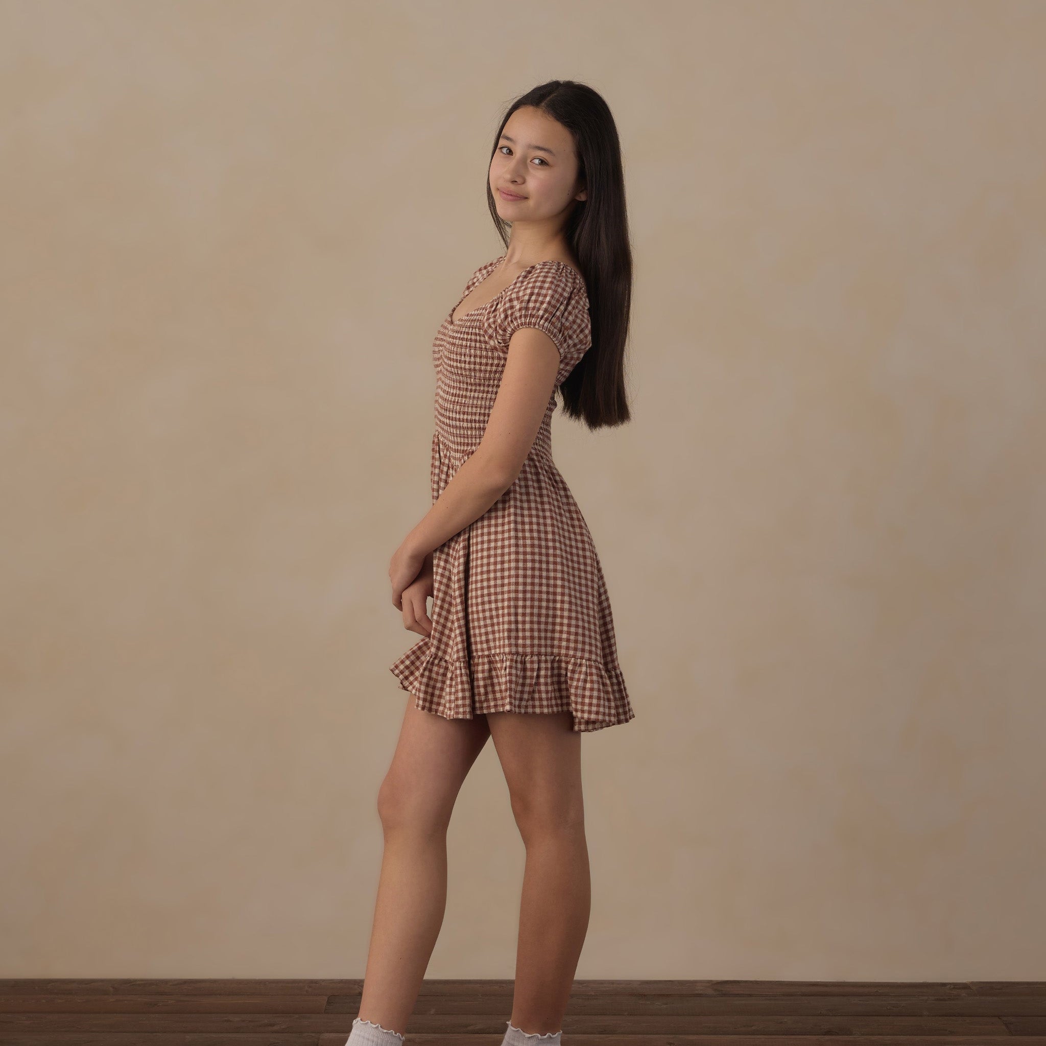 A young woman stands in profile wearing a short brown and beige gingham dress with puff sleeves, paired with white socks and black loafers, against a warm beige textured wall and dark wood floor.