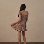 A young girl is shown from behind wearing a brown and beige checkered mini dress with puff sleeves, standing on a wooden floor against a textured beige wall.
