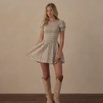 A woman stands in a studio wearing a short-sleeve fit-and-flare dress with a beige and white floral pattern, paired with light tan cowboy boots, against a warm beige wall and dark wood floor.