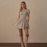 A woman stands in a studio wearing a short-sleeve fit-and-flare dress with a beige and white floral pattern, paired with light tan cowboy boots, against a warm beige wall and dark wood floor.